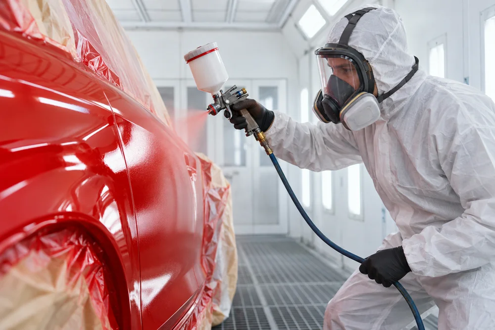 Illustration of a technician refinishing a red vehicle panel in a spray booth.